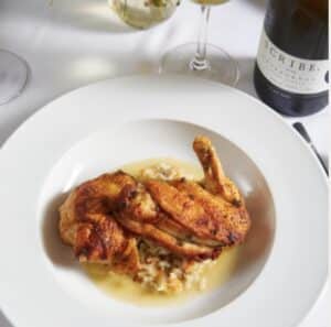 Roasted chicken on a bed of yellow rice, served on a white plate with a bottle of red wine of to the side.